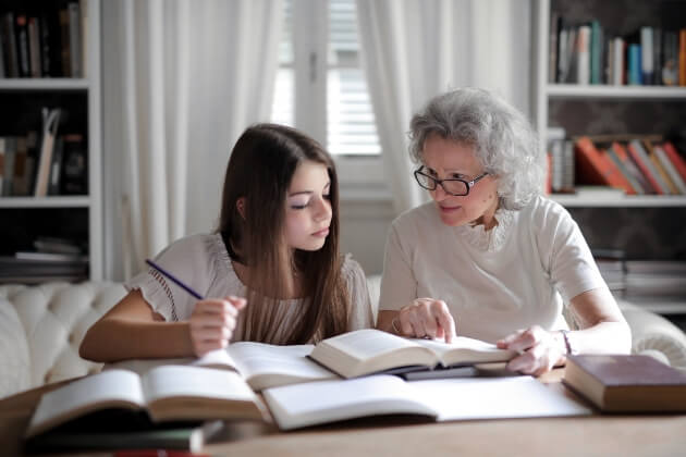A grandmother sitting at a table with several open books, helping her granddaughter with homework.