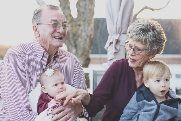 Grandparents sitting for a semi-formal photo outdoors with their infant and toddler grandchildren.