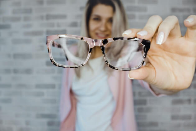 A pair of eyeglasses in the foreground being held out by a woman in the background.